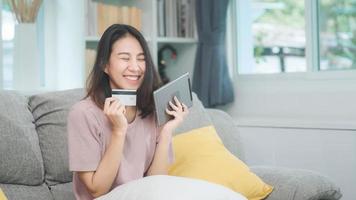 Young smiling Asian woman using tablet buying online shopping by credit card while lying on sofa when relax in living room at home. Lifestyle latin and hispanic ethnicity women at house concept. photo
