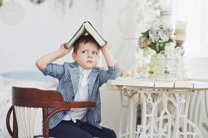 Photo of diligent schoolboy with book on his head doing homework. The schoolboy is tired of doing homework