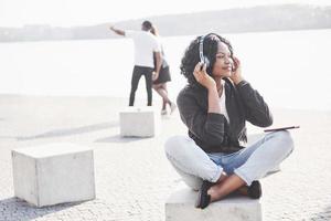 Retrato de una hermosa joven bastante afroamericana sentada en la playa o el lago y escuchando música en sus auriculares foto