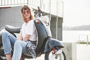 Retrato de una hermosa chica hipster sentada en un scooter retro negro, sonriendo posando y disfrutando del cálido sol primaveral. foto