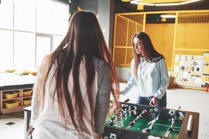 Two beautiful twin girls play table football and have fun. One of the sisters holds a toy ball in his hand and shows the tongue photo