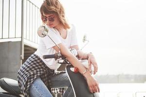 Portrait of a beautiful girl hipster sitting on a black retro scooter, smiling posing and enjoy the warm spring sunshine. photo