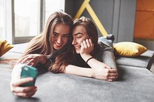 dos lindas hermanas gemelas sonrientes sosteniendo smartphone y haciendo selfie. las niñas se acuestan en el sofá posando y alegría