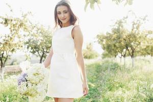 A beautiful young girl in a white light dress and a bouquet of summer flowers lays a fine day in the garden photo