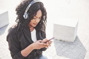 Retrato de una hermosa joven bastante afroamericana sentada en la playa o el lago y escuchando música en sus auriculares foto