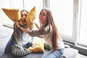 Two beautiful young twins sisters spending time together and are battling with pillows. Siblings having fun at home concept photo