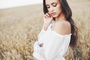 Sensible joven vestida de blanco posando en un campo de trigo dorado