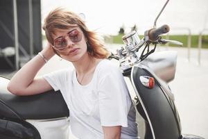 Portrait of a beautiful girl hipster sitting on a black retro scooter, smiling posing and enjoy the warm spring sunshine. photo