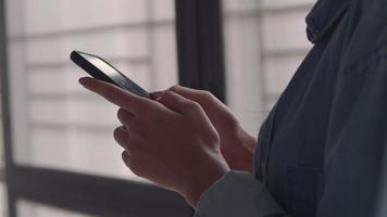 femme d'affaires asiatique tenant un téléphone portable et fait défiler le flux de médias sociaux dans un smartphone debout à côté de la fenêtre au bureau à domicile. video