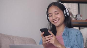 Asian woman listening to music in headphone use smartphone and laptop while sitting sofa in living room at home.
