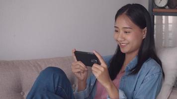 Asiatische Frau, die Videos auf dem Handy ansieht, die soziale Medien durchsucht, während sie zu Hause auf dem Sofa im Wohnzimmer sitzt.