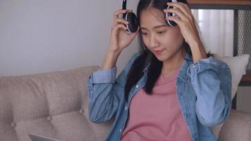 Mujer asiática escuchando música con auriculares, use un teléfono inteligente y una computadora portátil mientras está sentado en el sofá en la sala de estar en casa. video