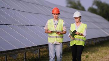 Two engineers are discussing drawings for the project. They are standing at a solar panel station. The female engineer teaches a young specialist who is undergoing practice video