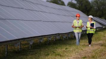 Two engineers are discussing drawings for the project. They are standing at a solar panel station. The female engineer teaches a young specialist who is undergoing practice video