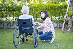 Ayude a la anciana asiática mayor o anciana en silla de ruedas y con una mascarilla para proteger la seguridad de la infección por el coronavirus covid-19 en el parque. foto