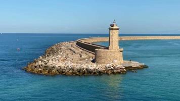 veduta aerea di un faro alto e robusto con rocce ai margini di un lungo porto sinuoso in mezzo all'ampio mare blu durante una luminosa giornata di sole, che ruota lentamente verso destra. video