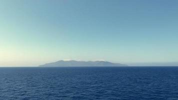 Wide angle of the vast blue sea gently waving during a calm and bright sunny day with an island visible above the horizon, panning to the left.