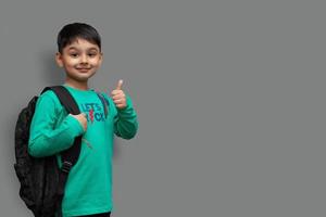 Happy smiling boy with thumb up is going to school. Child with school bag and books. Kid indoors on a background. Back to school.