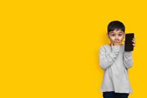 Sonrisa y conmocionado niño chico en camisa gris verde pantalla en blanco del teléfono móvil sobre fondo amarillo espacio de copia