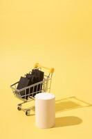 Podio simulado vacío o pedestal y carrito de supermercado en miniatura con bolsas de compras negras en venta de viernes negro sobre fondo amarillo