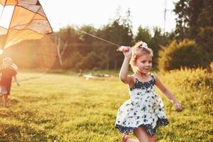 Linda niña con cabello largo corriendo con cometa en el campo en un día soleado de verano