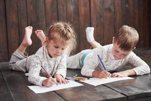 los niños se acuestan en el suelo en pijama y dibujan con lápices. lindo niño pintando con lápices foto