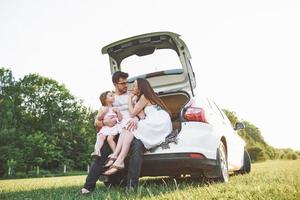 Pretty young married couple and their daughter are resting in the nature. The mother father and little girl are sitting on open car boot photo