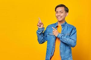 Cheerful young asian man pointing two fingers aside at copy space isolated over yellow background photo