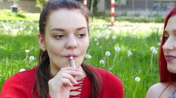 Dos hermosas chicas fumando cigarrillos electrónicos iqos en el parque de verano video