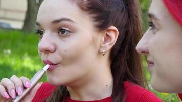 Zwei schöne Mädchen rauchen elektronische iqos-Zigarette im Sommerpark video