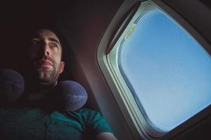 Hombre pensativo con asiento de almohada para el cuello por la ventana de un avión foto