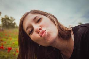 Mujer joven haciendo beso de cara de pato mientras toma una foto selfie con su teléfono inteligente o cámara en el campo de amapolas rojas