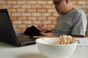 Thai male worker busy working with laptop, use chopsticks to hastily eat instant noodles during office lunch's break, because quick, tasty, and cheap. Over time Asian fast food, unhealthy lifestyle. photo