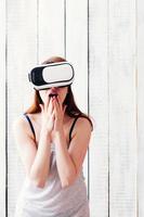 Chica con gafas de realidad virtual, emocionada, sosteniendo sus manos en la cara, fondo blanco de madera foto