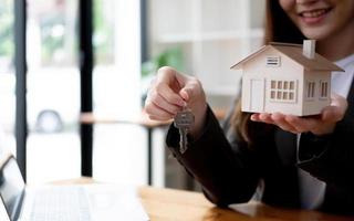 Attractive young female house realtor or real estate agent holding house keys chain and house model. blurred background. photo