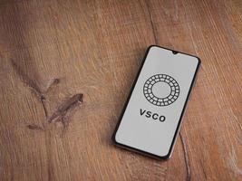 VSCO - Photo and Video Editor app launch screen with logo on the display of a black mobile smartphone on wooden background