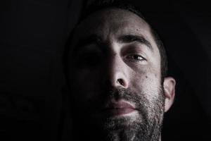 Dramatic portrait of a young man half expressionless face in deep shadow looking at camera isolated on black background photo