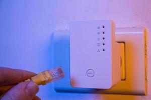 Man insert ethernet cable into WiFi extender device which is in electrical socket on the wall photo