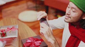 homens asiáticos que pegam uma caixa de foto-presente no smartphone decoram a árvore de Natal que comemora o ano novo na sala de estar em casa.