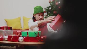 homens asiáticos verificando a caixa de presente decoram a árvore de natal comemora o ano novo na sala de estar em casa. video