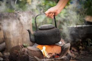 Cerrar mano sujetando hervir agua vieja tetera en el fuego con una estufa de carbón en el fondo borroso