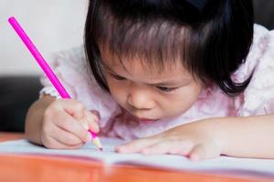 linda chica asiática está coloreando una madera de color rosa en un libro. niño miró de cerca. concepto de postura sentada y problema con los ojos. foto