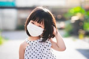 Girl smiled sweetly, showing thoughtful expression. Happy child put their hand on their heads. Little kid wearing white medical face mask. Children 4-5 years old wear white shirt with black polka dots photo