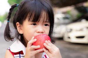 Head short of cute girl is biting at a red apple. Children eat fruit. Asian girl uses two hands to holding an apple. Little child is 3 and a half years old.