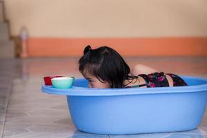 Active girl play water in blue plastic basin. Child looks at the heights of the two glasses. Baby are happy to enjoy summer or spring. A wet kids in black bathing suit. Cute Asian children 3 years old photo