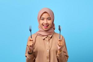 Retrato de mujer asiática joven sonriente mantenga tenedor y cuchara sobre fondo azul. foto