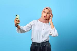 Retrato de mujer asiática joven conmocionada con la boca abierta mediante teléfono móvil sobre fondo azul. foto
