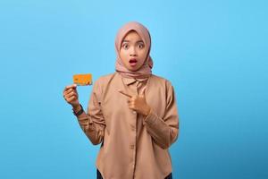 Retrato de mujer asiática joven sorprendida apuntando con tarjeta de crédito sobre fondo azul.