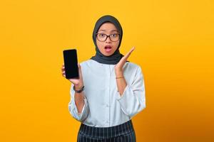Sorprendido joven asiática mostrando la pantalla en blanco del teléfono móvil aislado sobre fondo amarillo
