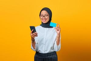 Alegre joven asiática sosteniendo teléfono móvil y tarjeta de crédito sobre fondo amarillo